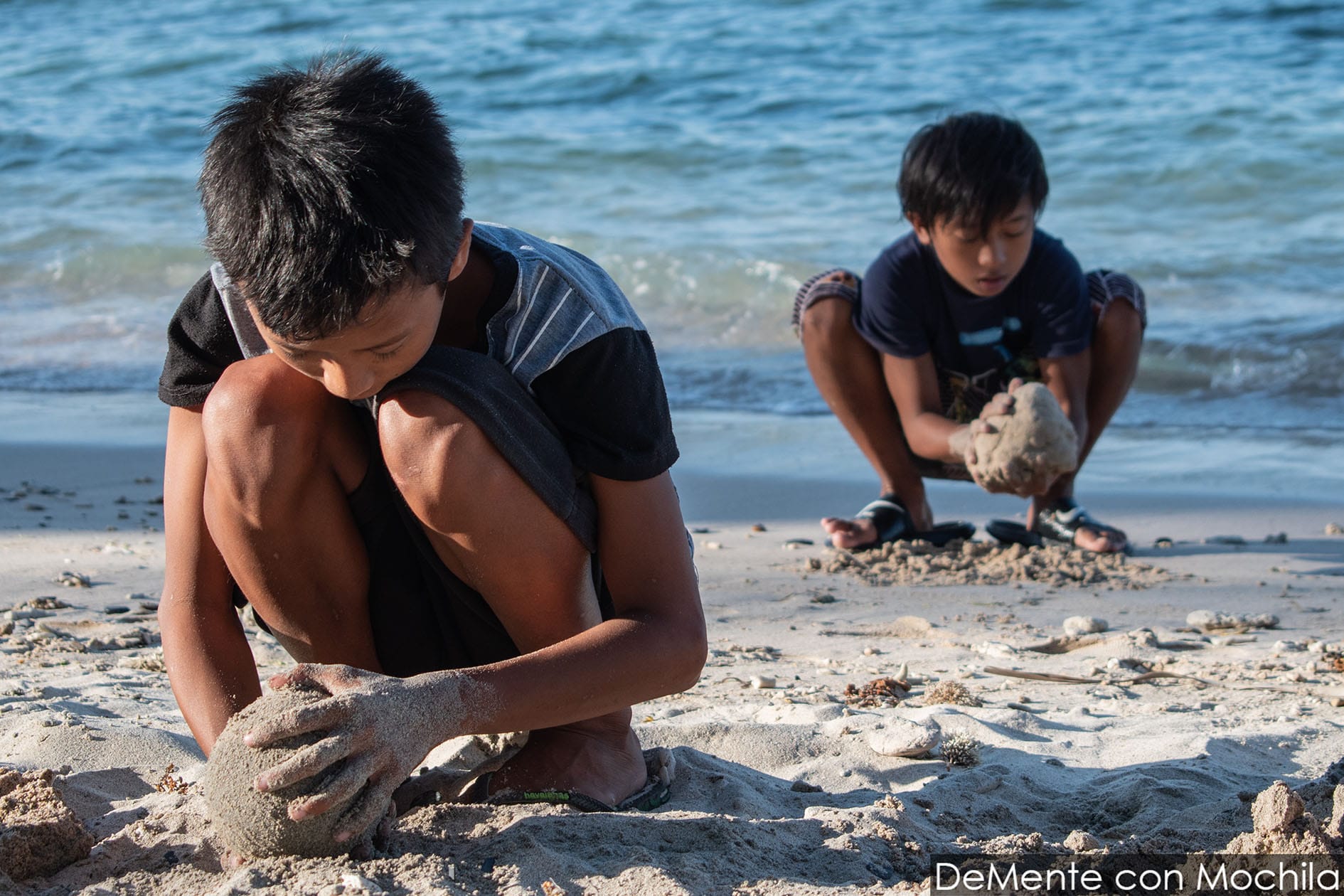 Dos niños arman bolas de arena: uno sobre la orillay el otro en arena seca